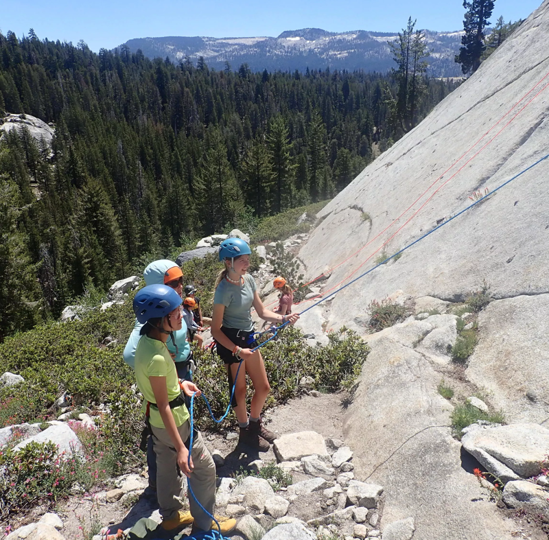 A group of young people are gathered at the base of a large rock face, preparing for or just finishing a rock climbing activity. They are wearing helmets and harnesses, and ropes are visible, suggesting they are engaged in belaying or climbing. The setting appears to be outdoors, with a forest and mountains in the background, indicating a natural climbing environment.